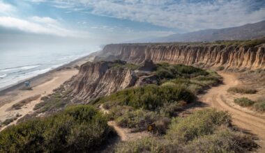A coastal landscape featuring cliffs, a sandy beach, and gentle waves under a cloudy sky.