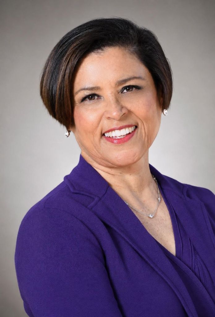 Professional headshot of a smiling woman with short dark hair, wearing a purple blazer and silver jewelry against a neutral gray background; warm, confident tone.