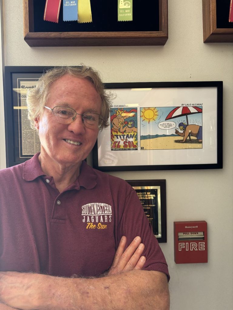 A smiling older man in a maroon Jaguars polo stands with arms folded beside framed vintage comic art, with a red fire alarm on the wall.