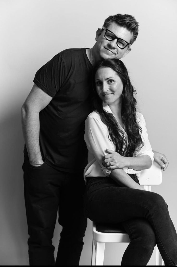 Black-and-white studio portrait of a smiling couple: a man in glasses leans over a seated woman in a white blouse, conveying warmth and closeness.