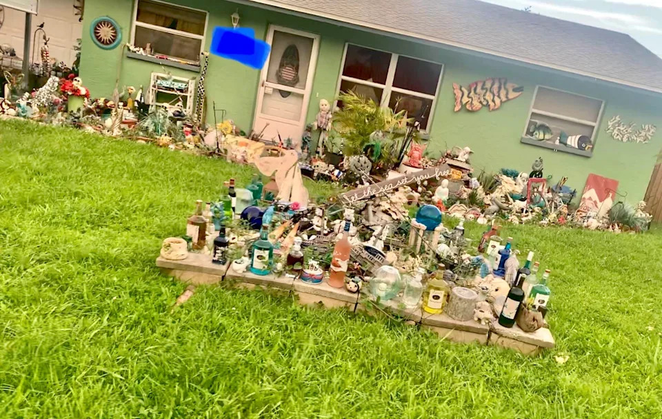 A front yard filled with numerous assorted decorations and bottles, with a green house in the background