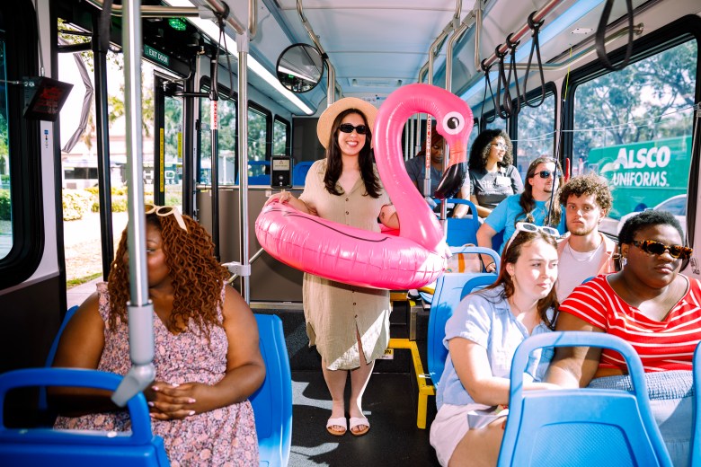 A smiling woman wearing a sun hat and sunglasses stands in the aisle of a public bus, holding a large, pink inflatable flamingo, surrounded by diverse passengers seated in blue seats.