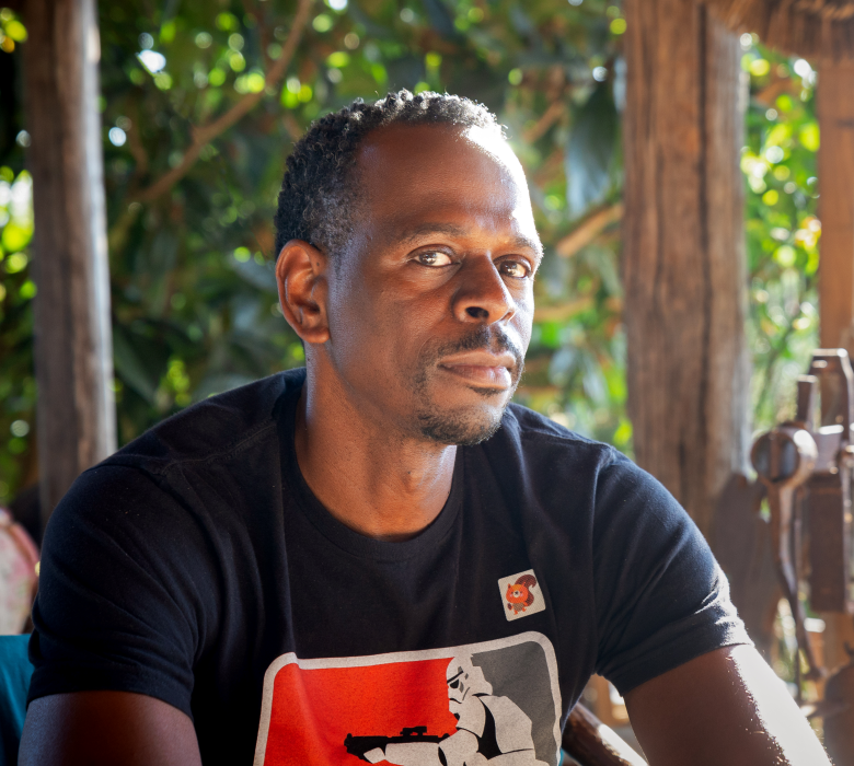 A man wearing a black T-shirt with a red and white graphic sits in a shaded outdoor area, looking directly at the camera with a calm, confident expression. Sunlight filters through greenery in the background.