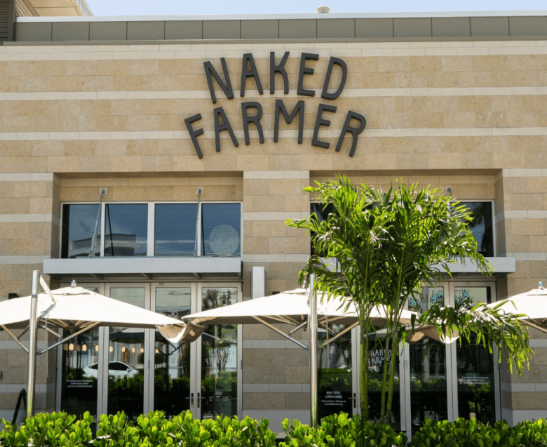 A straight-on, eye-level shot shows the exterior of a Naked Farmer restaurant. The building has a modern, tan-tiled facade with the name 'NAKED FARMER' in large, dark, sans-serif letters. In the foreground, an outdoor patio area features large white umbrellas, a small palm tree, and green bushes.