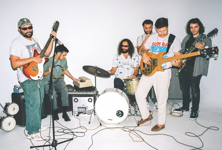 Seven musicians stand in a brightly lit studio, surrounded by instruments and tangled cords. Instruments visible include guitars, a drum kit, and a keyboard.