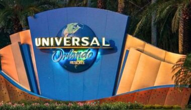The image shows the entrance sign for Universal Orlando Resort. The large, colorful sign boasts a globe design and is surrounded by palm trees and greenery, creating a vibrant welcome that feels like an invitation to join a community celebration.