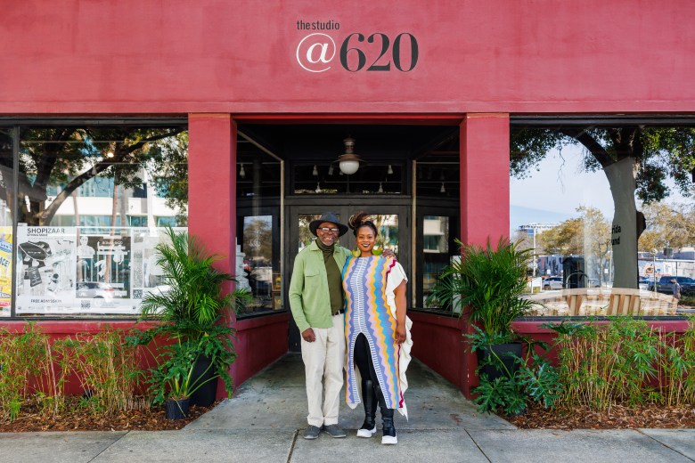 A man in a green shirt and hat, and a woman in a long, brightly colored striped dress, stand smiling in front of the red facade of "The Studio @ 620" venue.