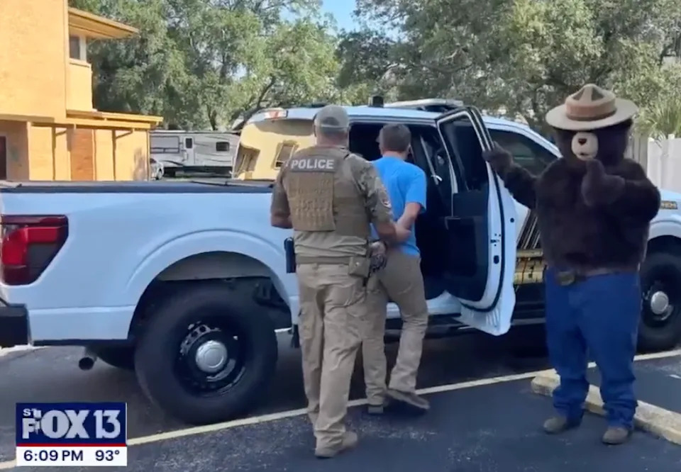 Police arrest a person near a pickup truck while someone in a bear costume stands nearby