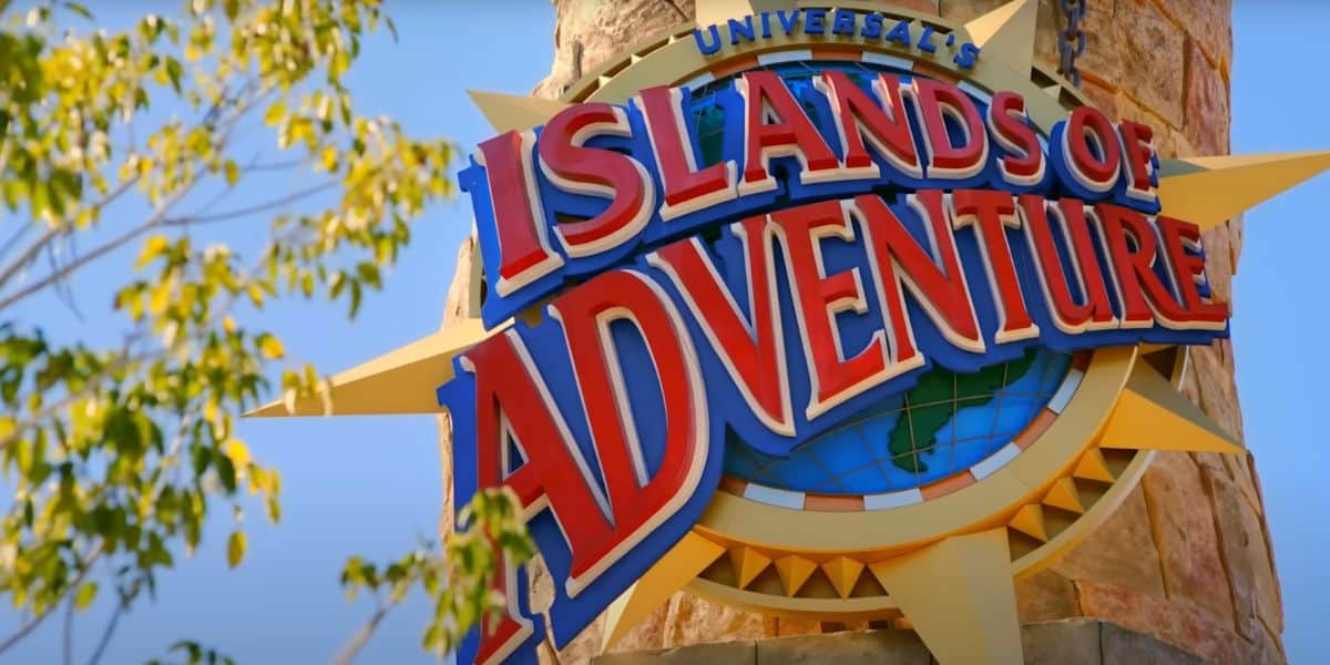 A colorful sign reading "Universal's Islands of Adventure" is mounted on a stone tower, with green leaves and a clear blue sky in the background, evoking excitement for attractions like Jurassic Park.