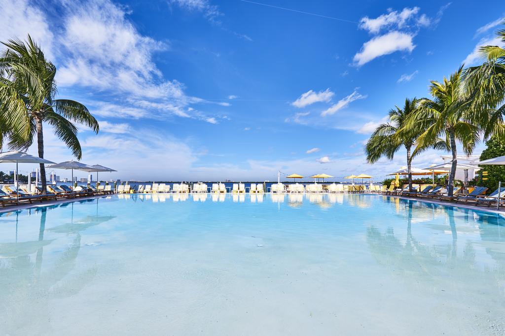 Infinity pool with clear water surrounded by palm trees and sun loungers, set against a bright blue sky with fluffy clouds.