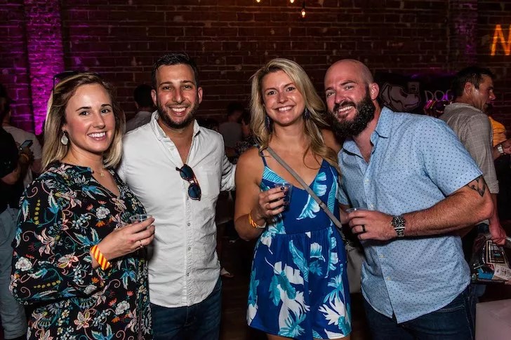 Four people—two women and two men—smile at the camera while holding drinks in a dimly lit venue with exposed brick walls and pink ambient lighting.