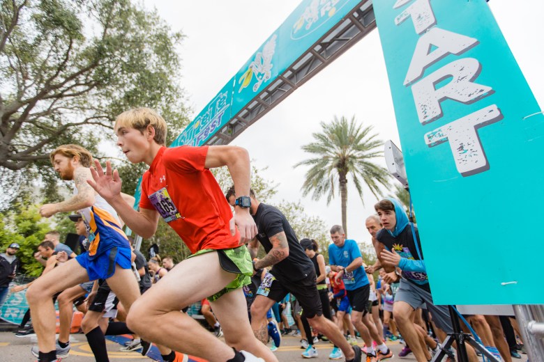 A low-angle action shot captures a group of runners as they burst forward from the starting line of a race. A large teal banner on the right reads "START" vertically in white letters. A young person in a red t-shirt and green shorts is in the foreground, mid-stride, surrounded by other competitors. A palm tree is visible in the background under an overcast sky.