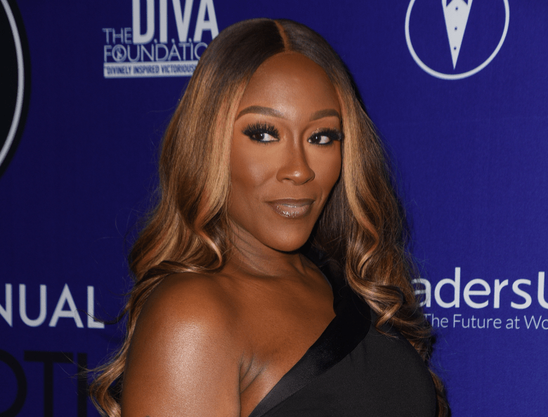 Close-up portrait of singer Coko from the group SWV, with long, wavy brown hair and dark eye makeup, posing at an event against a dark purple backdrop.