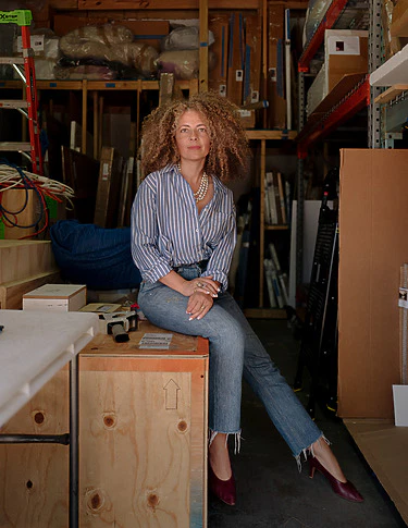 Person sitting casually on a wooden crate in a storage room, wearing a striped shirt and jeans, surrounded by various stored items.