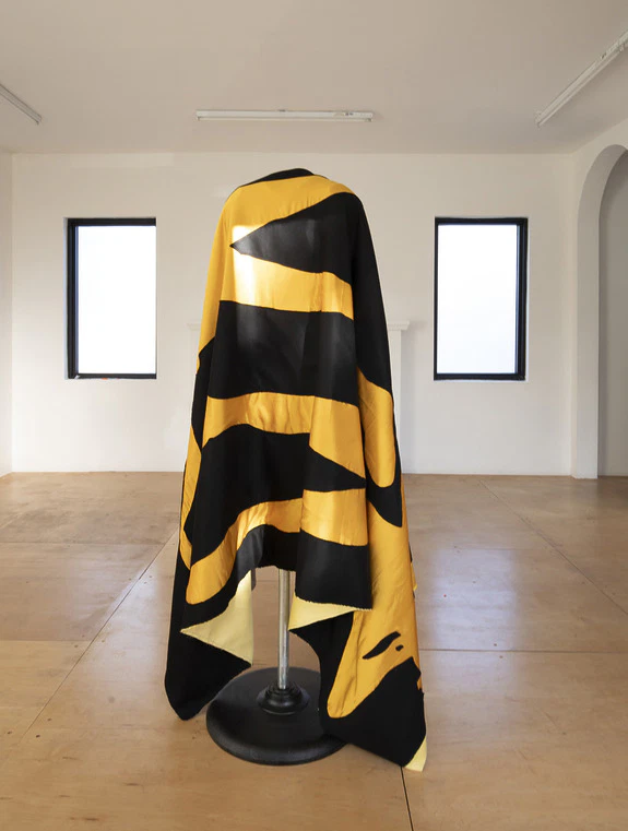 Black and yellow striped cloth draped over a mannequin in a minimalistic room with wooden floors and white walls.