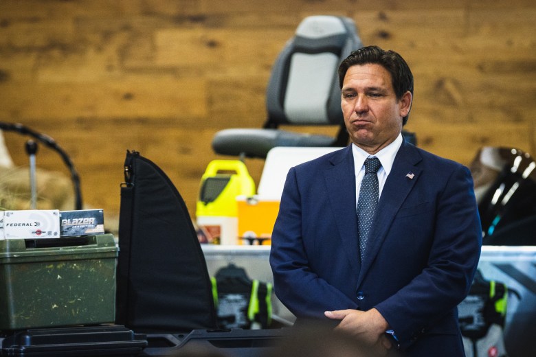 Governor Ron DeSantis, wearing a dark navy suit and tie, stands with a serious expression at an outdoor or indoor event. The background includes a wood-paneled wall and various items suggesting an outdoor theme, such as a boat seat, what appears to be a gun case or cover, and ammunition boxes.