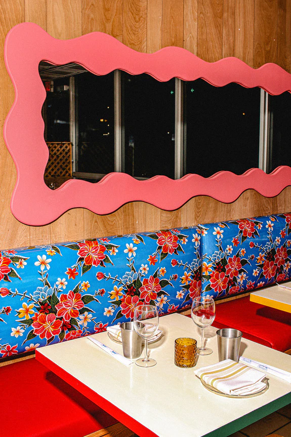 Colorful restaurant table setting with floral bench, pink wavy mirror, and neatly arranged silverware and glasses.