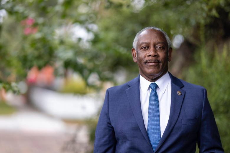 Outdoor portrait of Mayor Jerry Demings, wearing a dark blue suit and a patterned blue tie, smiling slightly against a blurred green, leafy background.