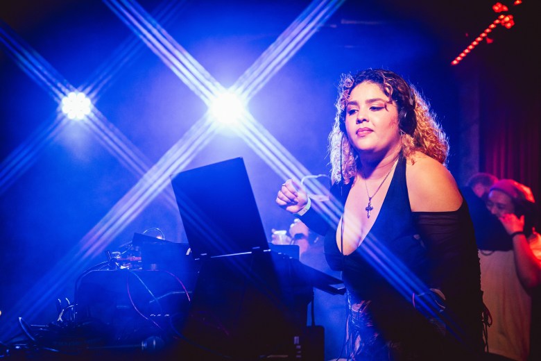 DJ Milla Killa performs on stage, wearing headphones and a black top, illuminated by bright, crossed blue spotlights while working at a laptop and mixer.