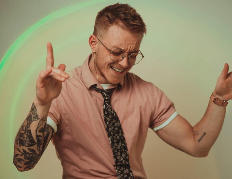 A medium shot of a stylish young man with short reddish-brown hair, round glasses, and a long chain earring, looking down and smiling brightly as if dancing. He wears a pink short-sleeved shirt and a black floral tie. His arms are raised, and he has prominent tattoos on both forearms. A soft arc of green light glows on the plain background behind him.