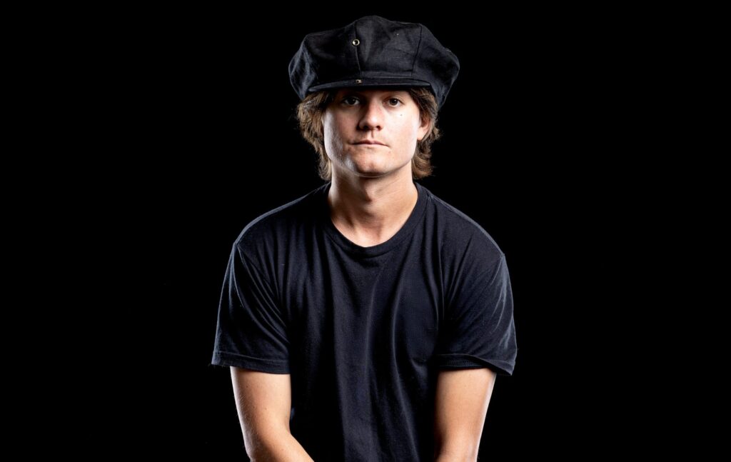 A studio portrait of musician Robin Taylor Zander wearing a black t-shirt and a dark cap, looking directly at the camera against a black background.