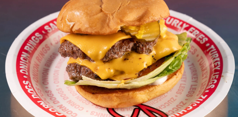 A juicy double cheeseburger served on a disposable white paper plate printed with red text like "ONION RINGS" and "CORN DOGS." The burger features two beef patties, melted yellow cheese, pickles, and lettuce on a soft, toasted bun.