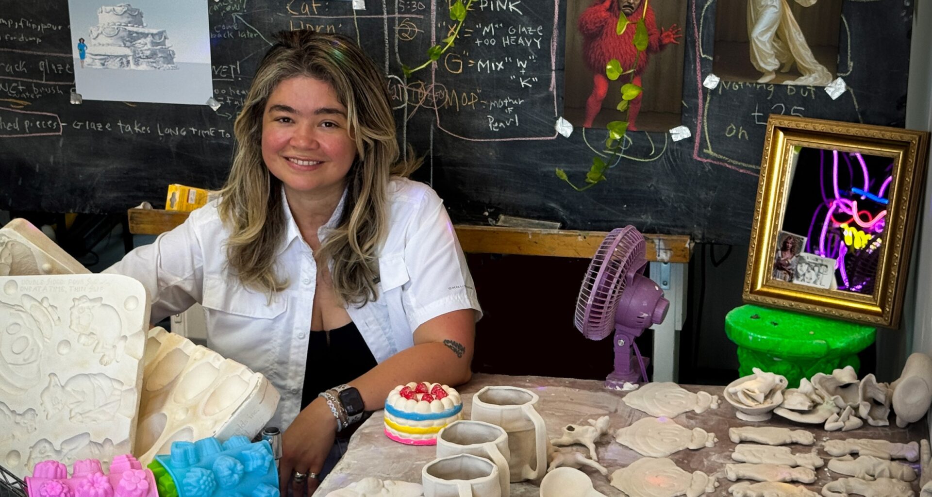 Photo of an artist in her studio. Behind her is a chalkboard with writing and posters. On the table beside her are clay scultures
