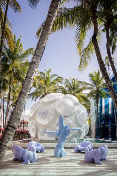 Sculpture garden featuring abstract blue figures and a geodesic dome surrounded by palm trees under a clear blue sky.