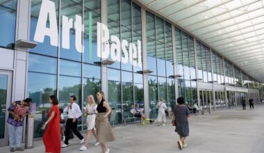 photo of people walking in front of the glass windows of a convention center with a sign on it reading, "Art Basel"