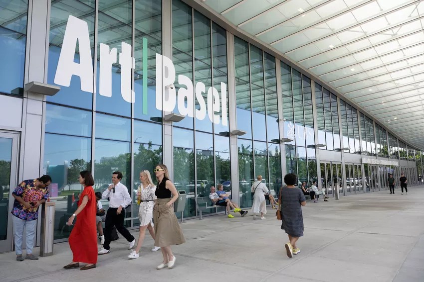photo of people walking in front of the glass windows of a convention center with a sign on it reading, "Art Basel"