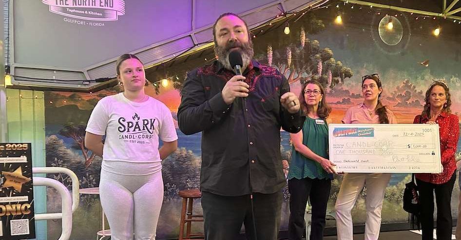 A man holding a mic, a girl standing behind him in white and 4 people holding a giant check.