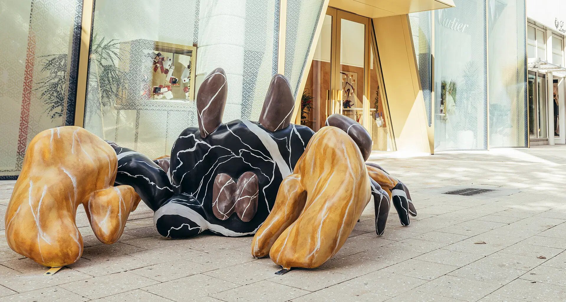 Abstract sculpture with black, white, and brown forms on a city sidewalk near a modern building entrance.