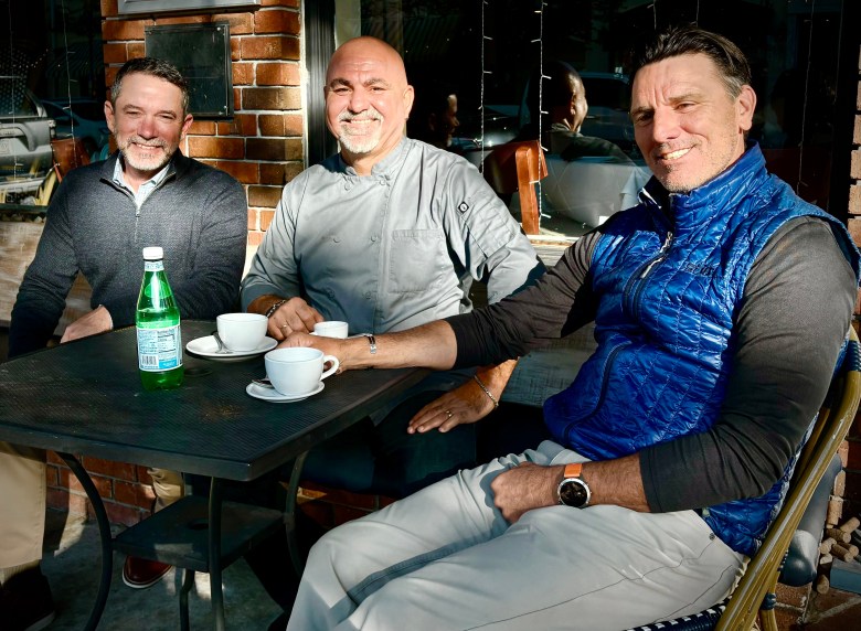 three men sit at an outside cafe table in the sun