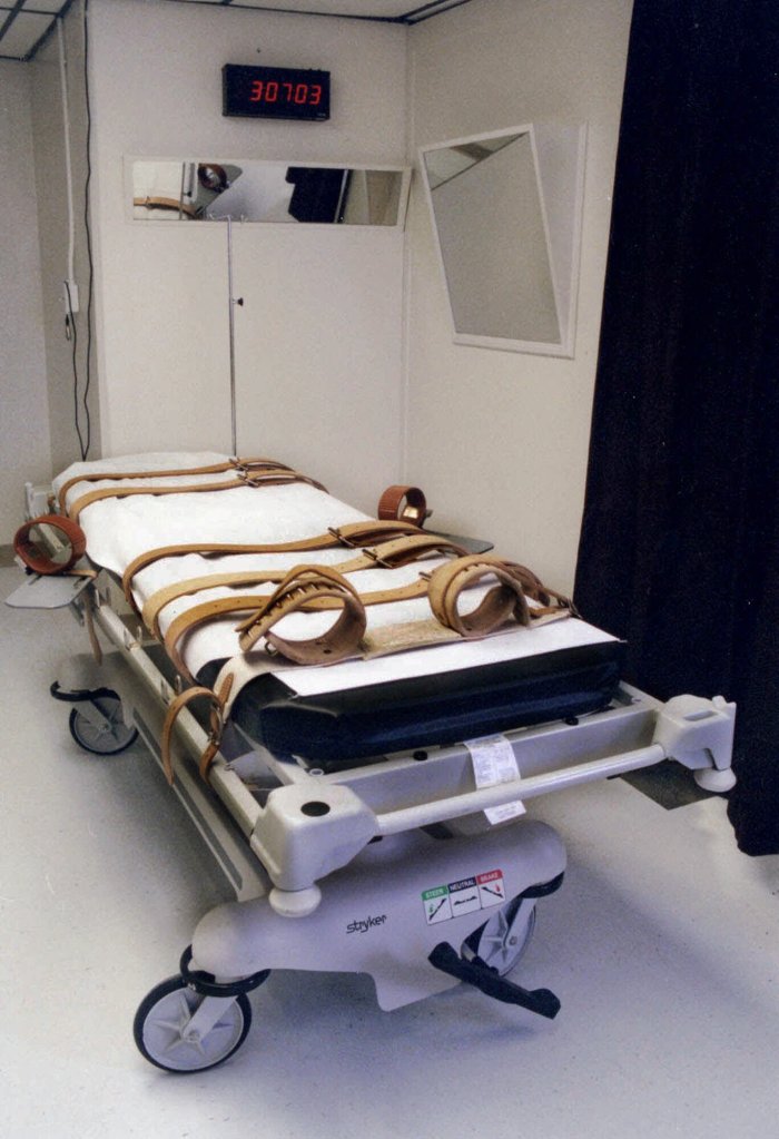 A gurney with leather straps, used for lethal injection, is in a room with a digital clock displaying "30703."