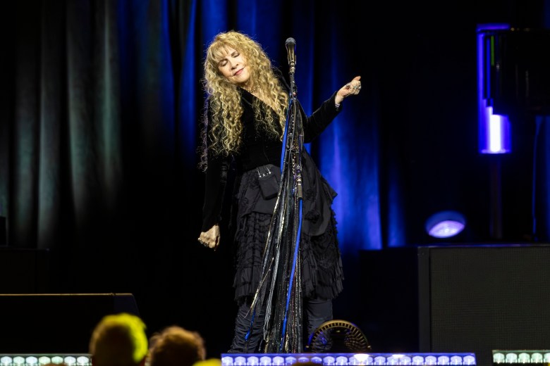 A performer with long curly blonde hair stands on stage wearing a flowing black outfit, eyes closed and hands raised slightly as she sings into a microphone draped with scarves. Blue and purple stage lighting illuminates the dark background.
