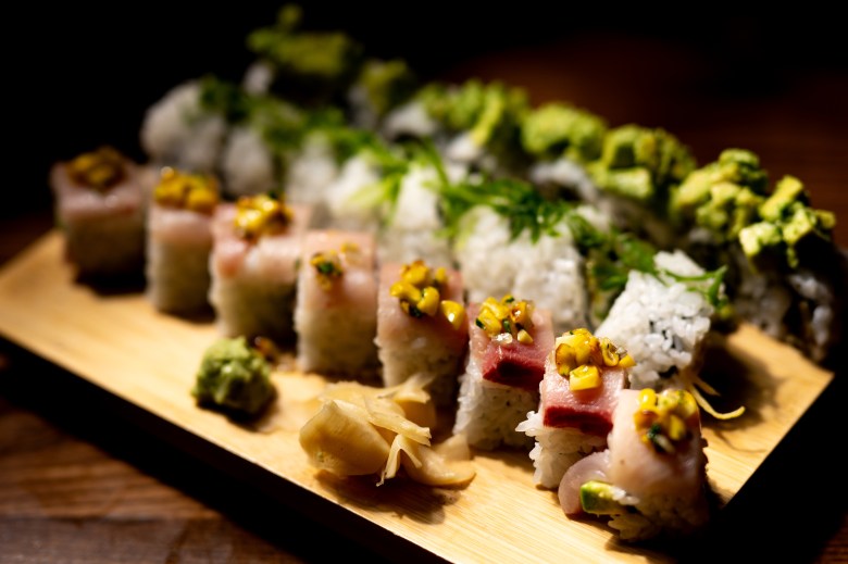 A rectangular wooden board displays several rows of sushi rolls, dramatically lit against a dark background. The front row features rolls topped with slices of pink raw fish and a yellow corn relish or garnish, while the back rows include rolls topped with avocado and herbs. A pile of pickled ginger and a dollop of wasabi sit on the board in the foreground.