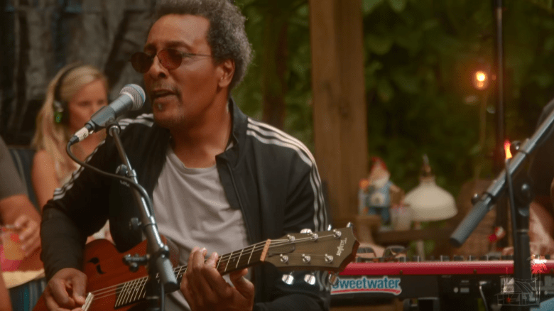 A close-up of a musician with salt-and-pepper hair and sunglasses, singing into a microphone while playing an acoustic guitar. They are wearing a black track jacket with white stripes on the sleeves over a grey t-shirt.