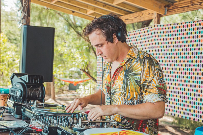 A side-profile photograph of a DJ with short brown hair, wearing black headphones and a vibrant, patterned tropical shirt. They are focused on mixing music using two turntables and a mixer on a wooden stage under a rustic pavilion at Pine Key, also known as Beer Can Island.