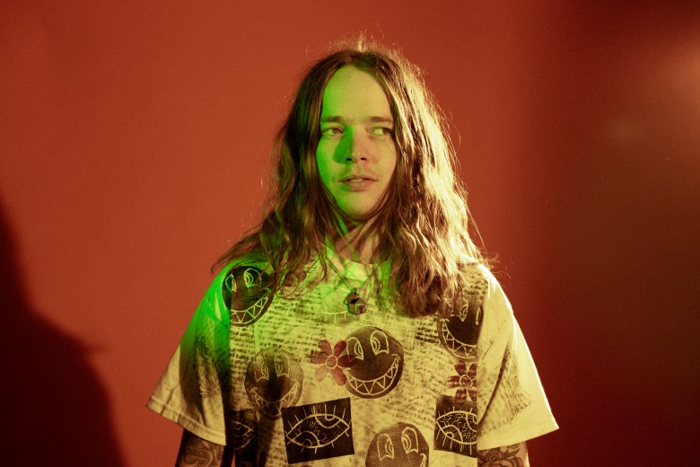 A person with shoulder-length wavy hair and a neutral expression looks off to the right. The subject is wearing a printed t-shirt featuring smiley faces and eyes, with tattoos visible on the arms, under harsh studio lighting.