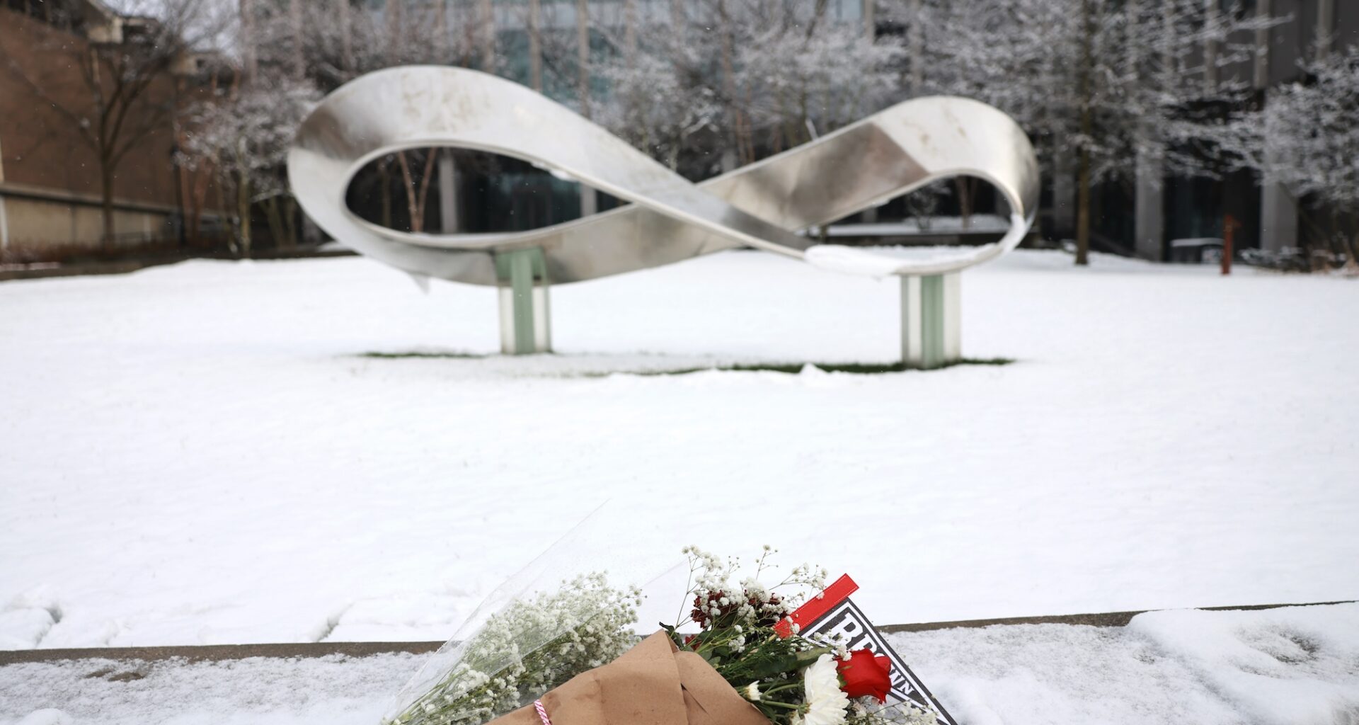 two bouquets of flowers placed atop a low stone wall dusted with snow. in the background, a large sculpture in the shape of the infinity symbol / möbius strip