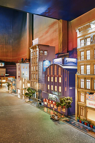 Miniature city street scene with colorful buildings, neon signs, small cars, and sunset backdrop creating a vibrant urban atmosphere.