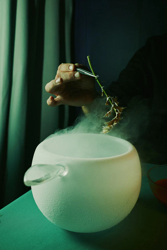 Chef delicately places herb into a smoking white ceramic bowl with tongs, creating a mystical and aromatic presentation.