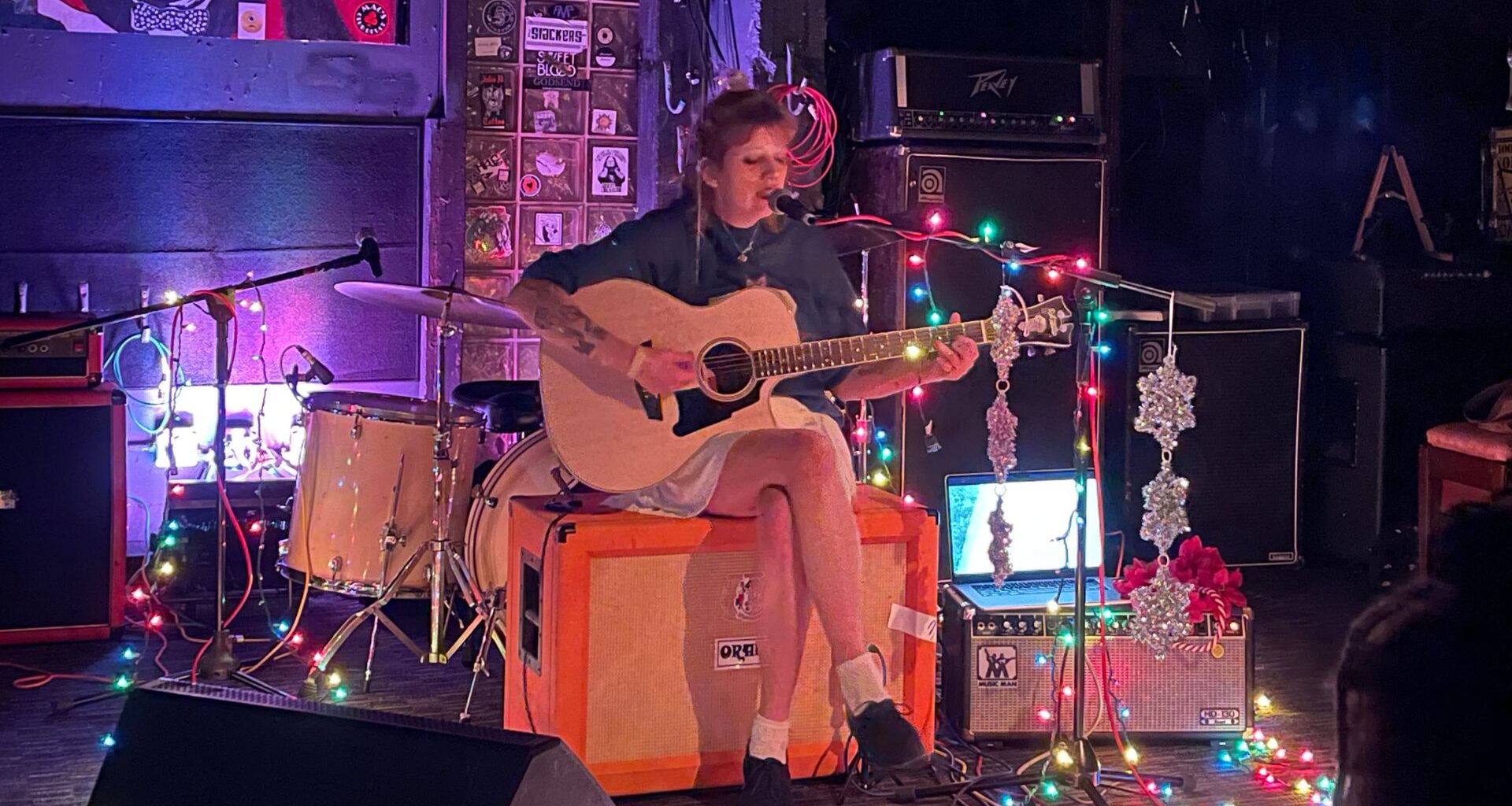 A photo of a woman playing a guitar sitting on an amplifier.