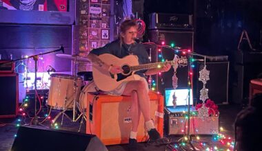 A photo of a woman playing a guitar sitting on an amplifier.