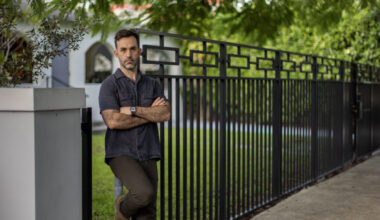 a man stands with folded arms leaning on a black iron fence