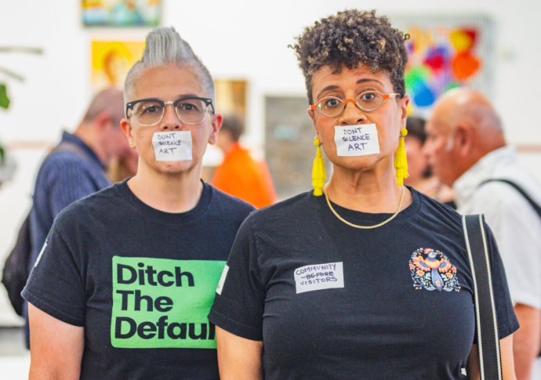 Two people stand side by side at an art event with tape over their mouths that reads “DON’T SILENCE ART.” The person on the left has short gray hair, glasses, and a black shirt with bold green text that says “Ditch The Default.” The person on the right wears bright yellow tassel earrings, glasses, and a black shirt with a patch design and a sticker that reads “COMMUNITY BEFORE VISITORS.” Blurred attendees and colorful artwork are visible in the background.