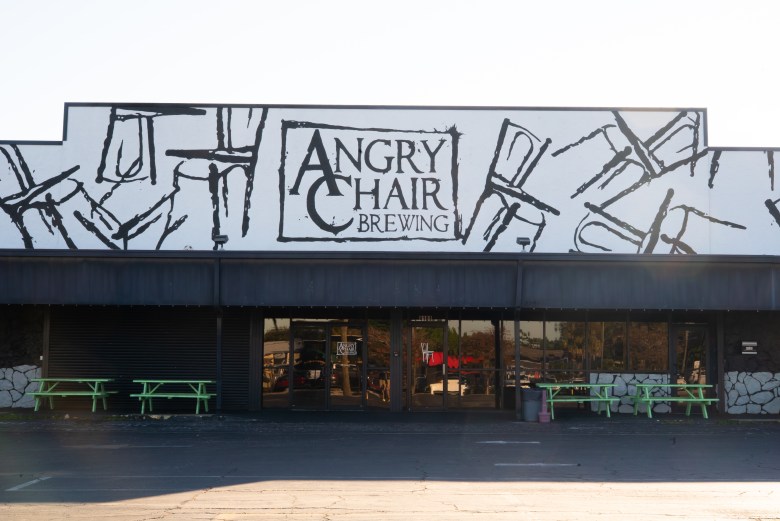 The exterior of the Angry Chair Brewing building features a distinctive mural above the awning, depicting abstract black sketches of chairs against a white background. Below the dark awning, the storefront has large tinted windows and glass entrance doors. Several bright green picnic tables are arranged on the pavement in front of the building.