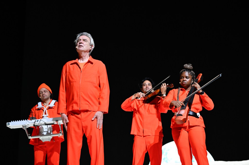 Four musicians standing on stage wearing orange jumpsuits. 