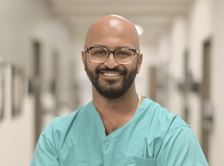 A professional portrait of an individual with a bald head and a full beard, wearing square-rimmed tortoiseshell glasses and teal-colored medical scrubs. The person is smiling and looking directly at the camera.