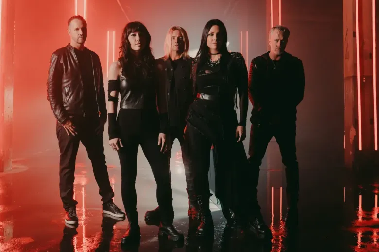 A full-length promotional photo of the rock band Evanescence, featuring five members standing in a dimly lit, warehouse-like space. The room is saturated with a dramatic red glow from vertical light tubes placed on either side of the band and behind them. The floor is reflective, mirroring the red light.